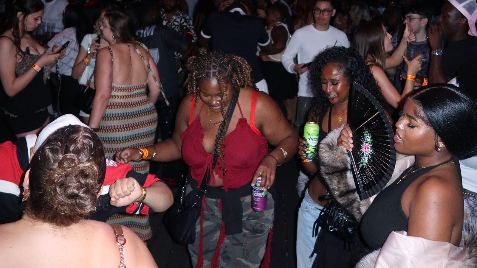 A group of women hold alcoholic drinks in their hands as they dance at a music event.