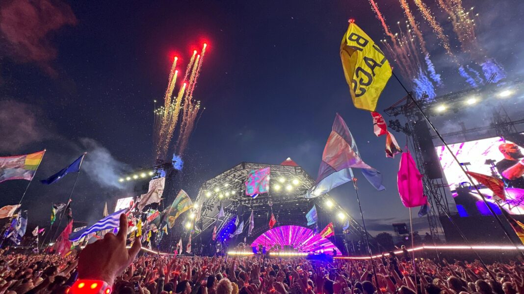 The Pyramid Stage at Glastonbury Festival. There is a huge crowd of people watching a performance. There are lots of flags waving above the audience and fireworks going off.