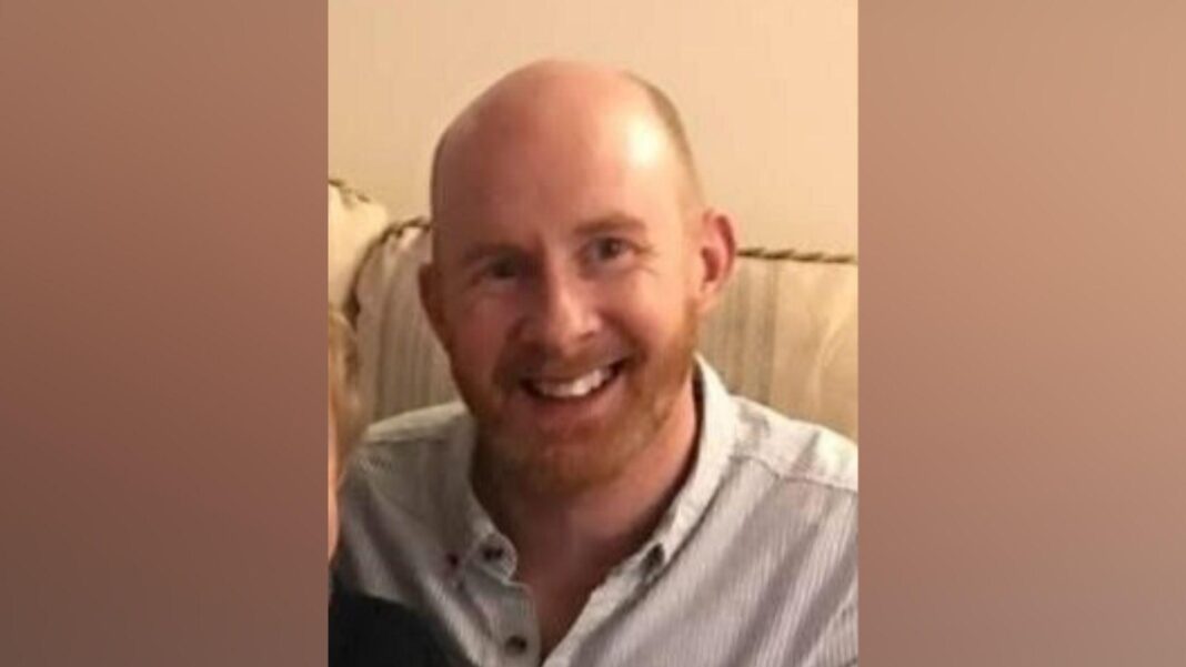 A man smiling at the camera in a head and shoulders image. He has a bald head with very short ginger hair at the sides and a ginger beard. He is wearing a pale blue shirt and sitting on a cream sofa.