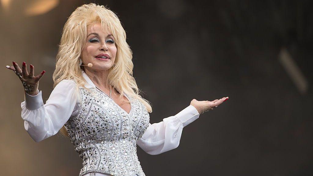 Dolly Parton in a white rhinestone top with blonde hair, and her arms outstretched.
