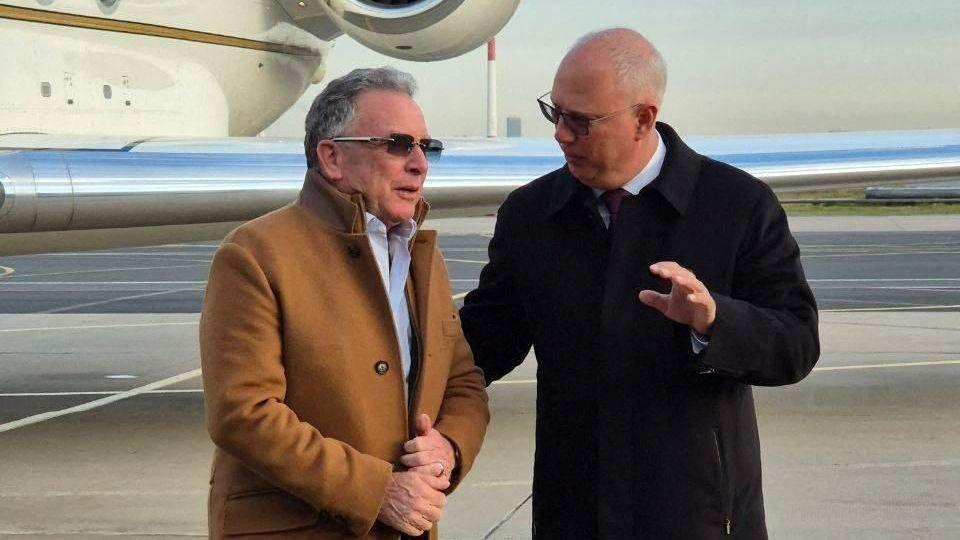 Witkoff, wearing a brown winter jacket, speaks to a man dressed in black, with a jet behind them.