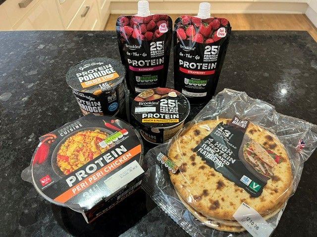 The image shows a peri peri chicken dinner, a porridge pot, 2 tall yoghurt pouches, 1 smaller yoghurt pot and a pack of flat breads - all labelled clearly with the word protein clearly displayed.