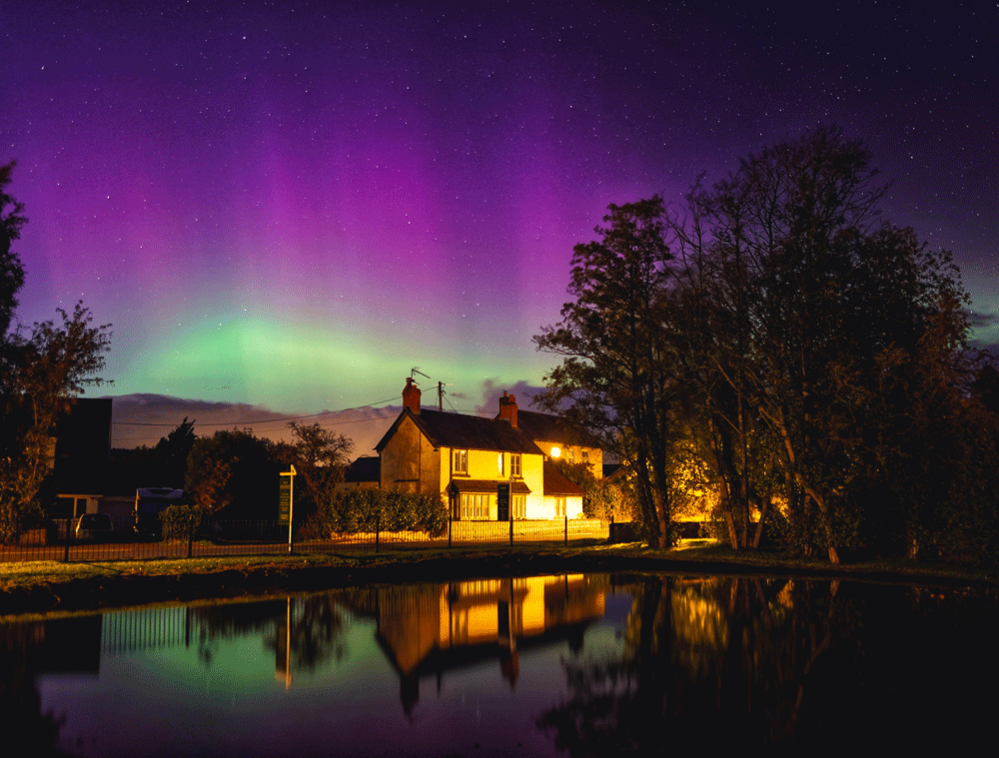 An image of the sky on 10 October during the evening. The Northern Lights can be seen in purple, pink and green hues in the sky and reflecting on a watery surface.