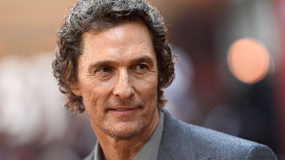 Matthew McConaughey is smiling at a camera. He has short brown curly hair, and is wearing a grey suit. This photo was taken at a screening for a film he stars in called "The Lost Bus”.