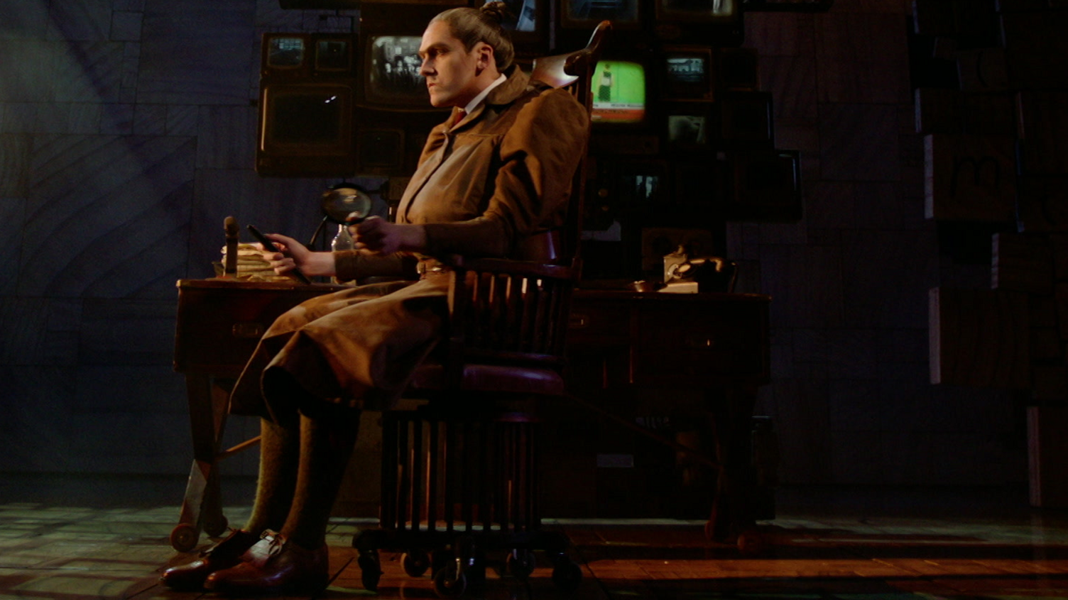 A stern female character sits in a chair on stage. They are wearing a brown leather-looking skirt and jacket with hair tied back in a bun.