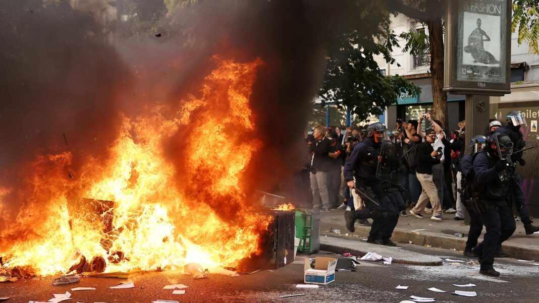 France halts use of stun grenades after boy’s hand blown off in student protests A large fire burns on a road in Paris beside which five officers in riot gear walk past a crowd observing the flames. There is debris strewn across the street.