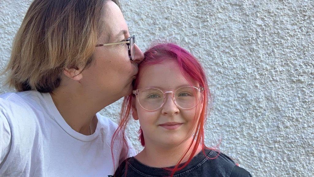 A woman with brown hair and glasses kisses her daughter who has dyed red hair and glasses on the side of her head.