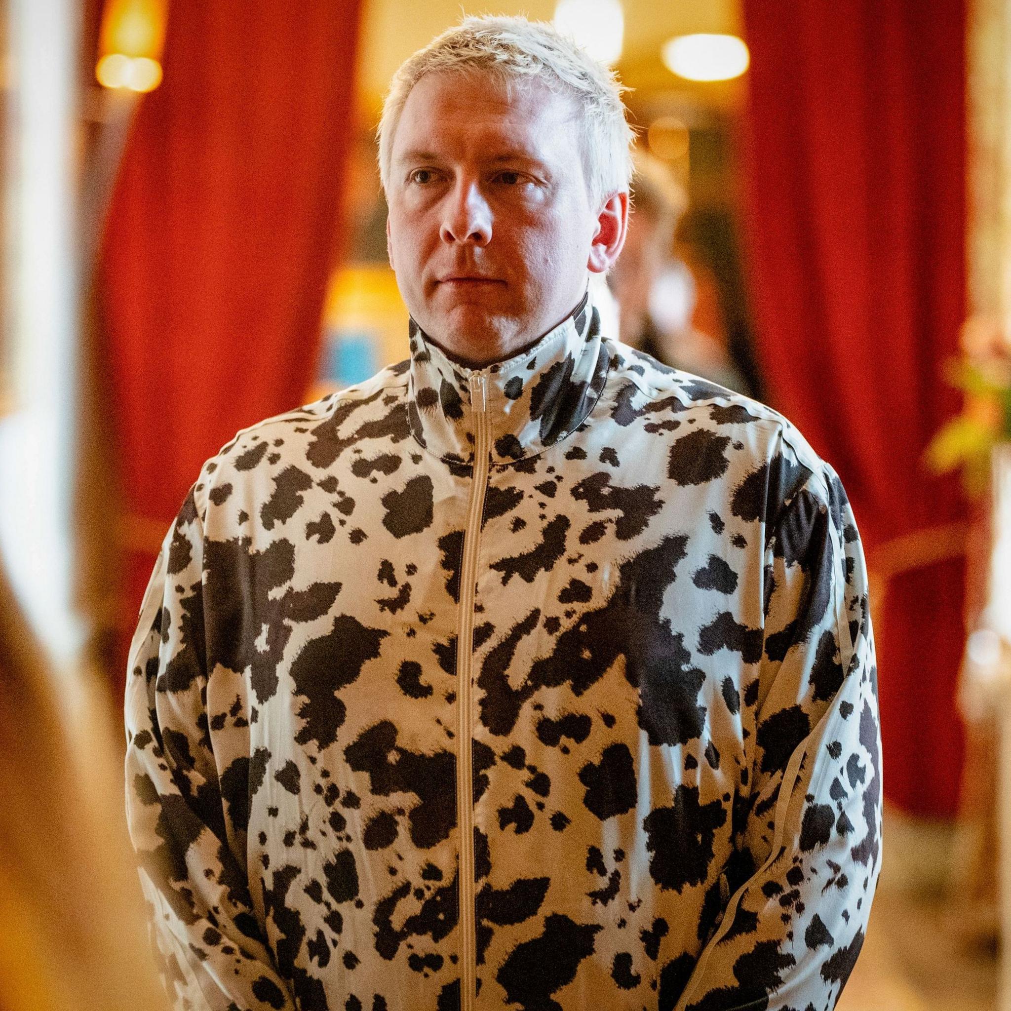 Joe Lycett standing in a zipped up white jacket with numerous black spots and splodges