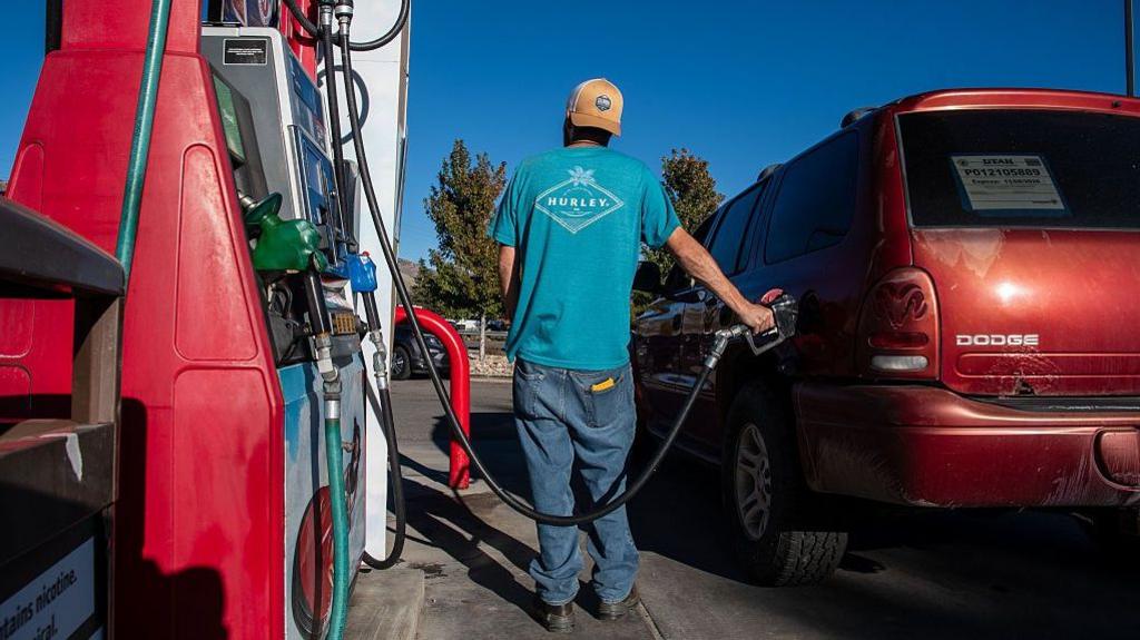 A customer wearing a backwards cap and a blue tshirt refuels a red vehicle at a gas station in Salt Lake City, Utah, United States on a clear, sunny day.