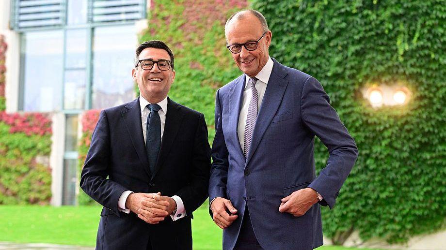 Andy Burnham smiles next to Friedrich Merz, both in navy blue suits, with green foliage in the background.