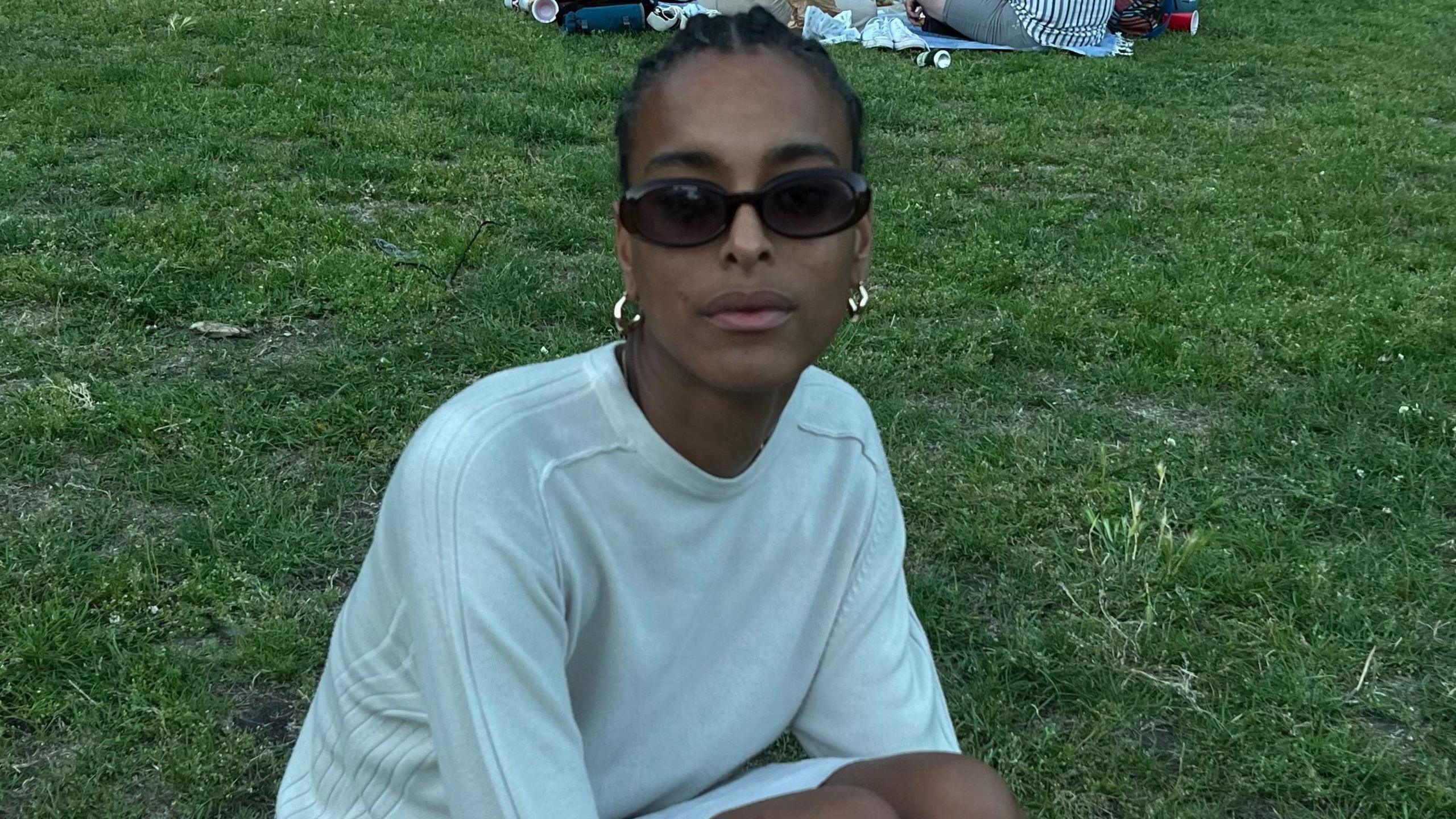 Summer wears a white jumper and sits on the grass. She has black hair braided back and wears sunglasses and gold earrings.