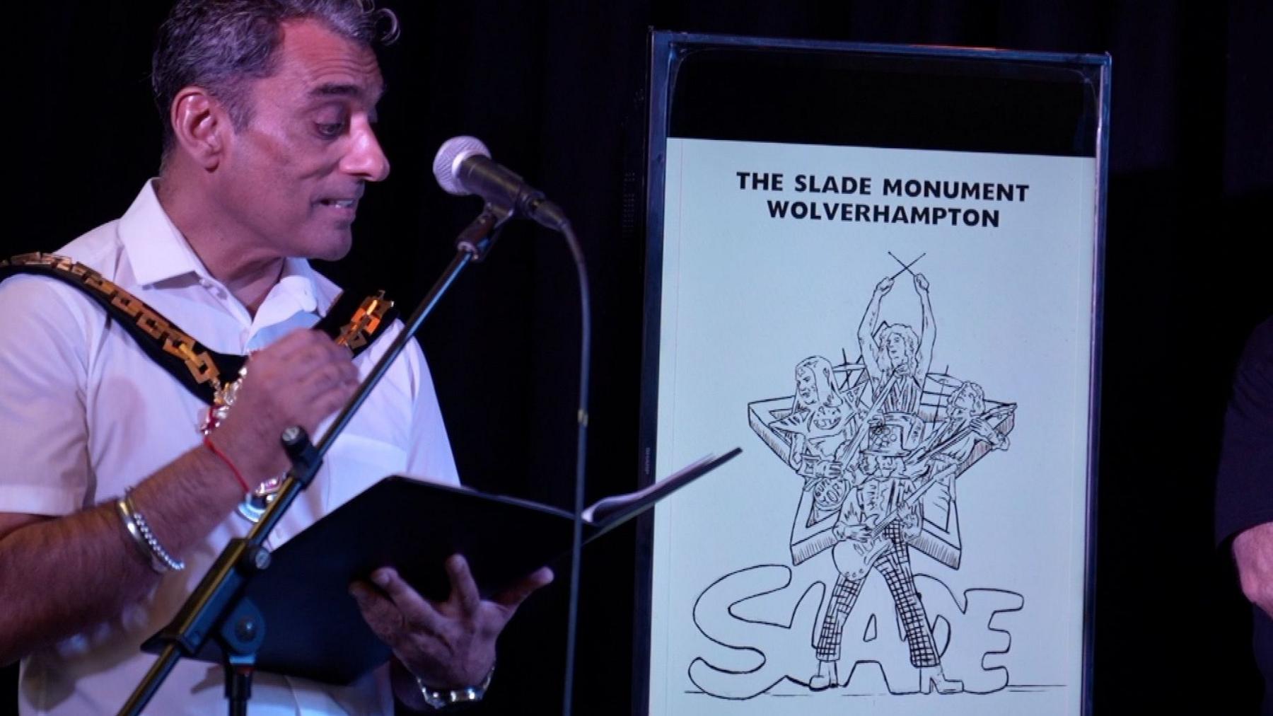 A man is wearing a white shirt and is standing in front of a microphone, next to a drawing of a monument.