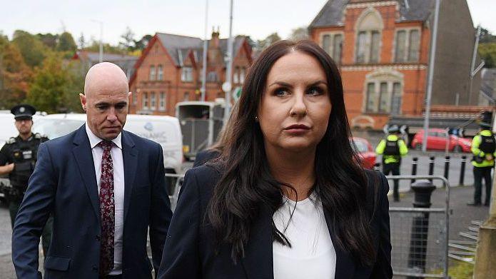 A woman with long brown hair. She is standing outside, and is wearing a white top and a dark blazer. In the background, there are a number of police officers.