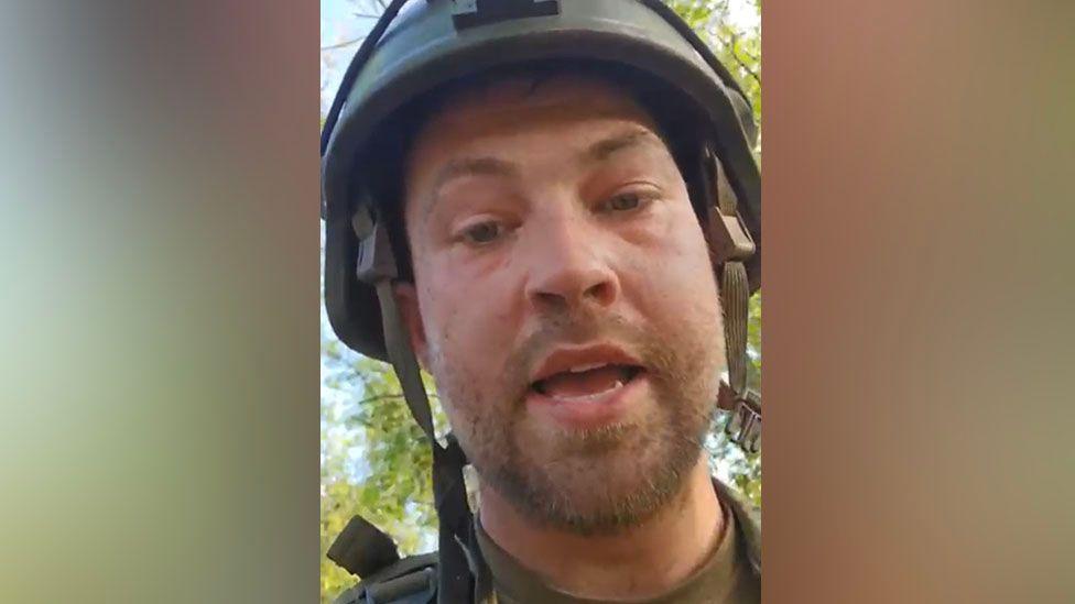 Nathan Stobbart is dressed in khaki army uniform and is wearing a flak helmet with the under-chin strap not done up. He has beard and moustache stubble and bags under his eyes and looks tired. He has his mouth open in mid-speech. Behind him are green, leafy trees and a pale blue sky.
