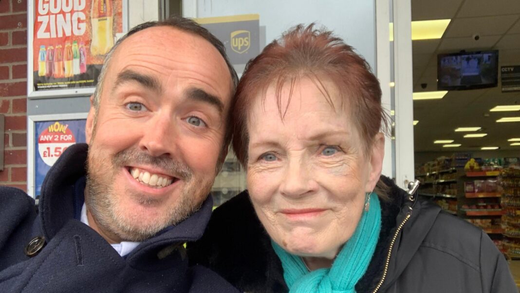 Matt Murray, wearing a navy blue coat and with a short trimmed beard and moustache, smiles for a selfie next to Anne Hughes, who had short red hair and a blue scarf