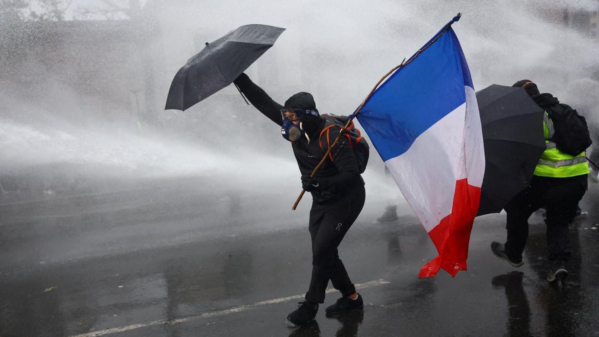 A masked prostester holding an umbrella and a French flag reacts as French police use a water canon amid clashes during an authorised demonstration organised by labour unions in Paris.