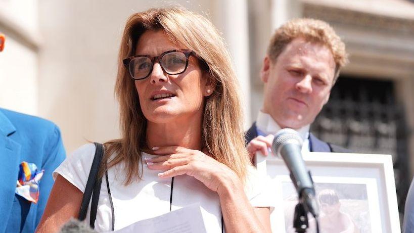 Ellen Roome, a woman with glasses and fair hair is stood behind a microphone with a framed photo of her son in the background.