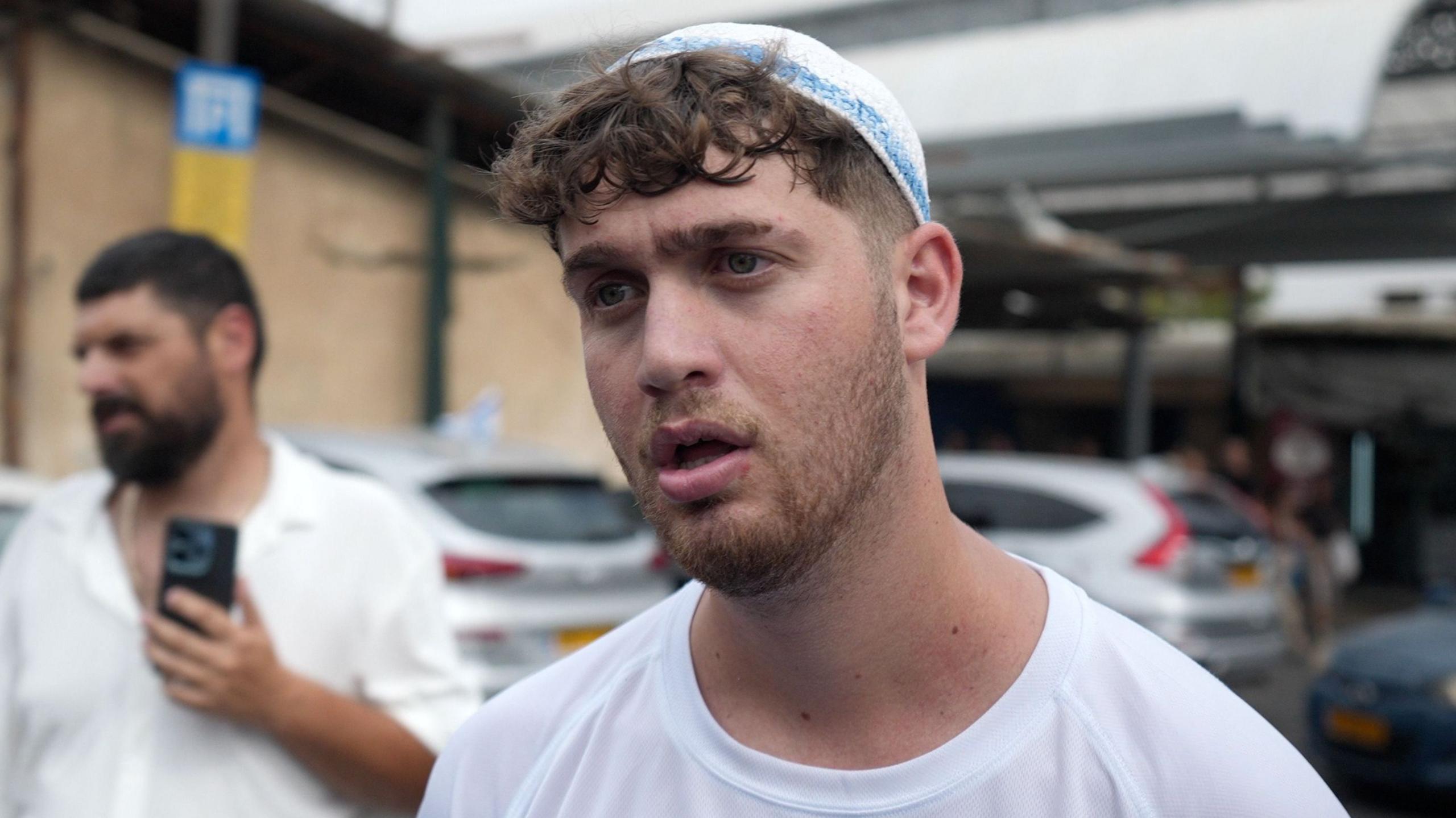 A man in a white t-shirt and a white skullcap, also known as kippah, talks to camera