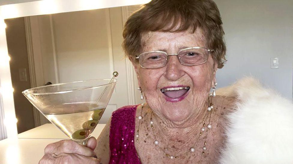 Ninety-six-year-old Grandma Droniak, holding a martini glass and wearing a pink sparkly top and a white fur shawl. She is standing in front of a dressing table with lights around the mirror.
