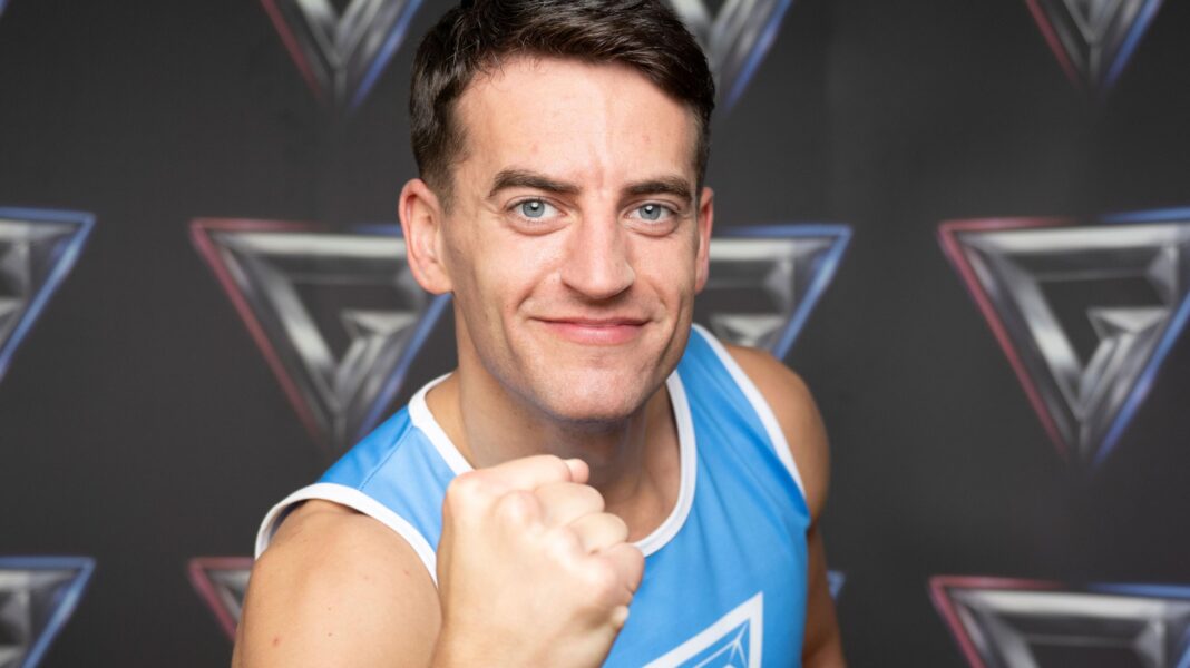 Josh McDonald smiles for the camera, while holding a raised fist. He is wearing a blue lycra jumpsuit, and standing in front of a backdrop with the Gladiators logo.
