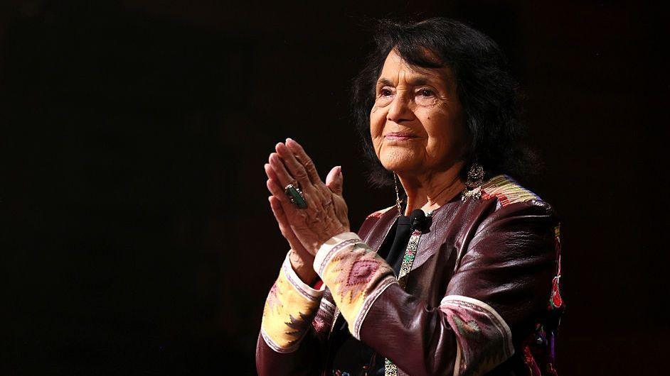 Dolores Huerta slightly turned to her right with her hands in a prayer position and a large turquoise ring on her right hand wears a brown leather jacket and long earrings