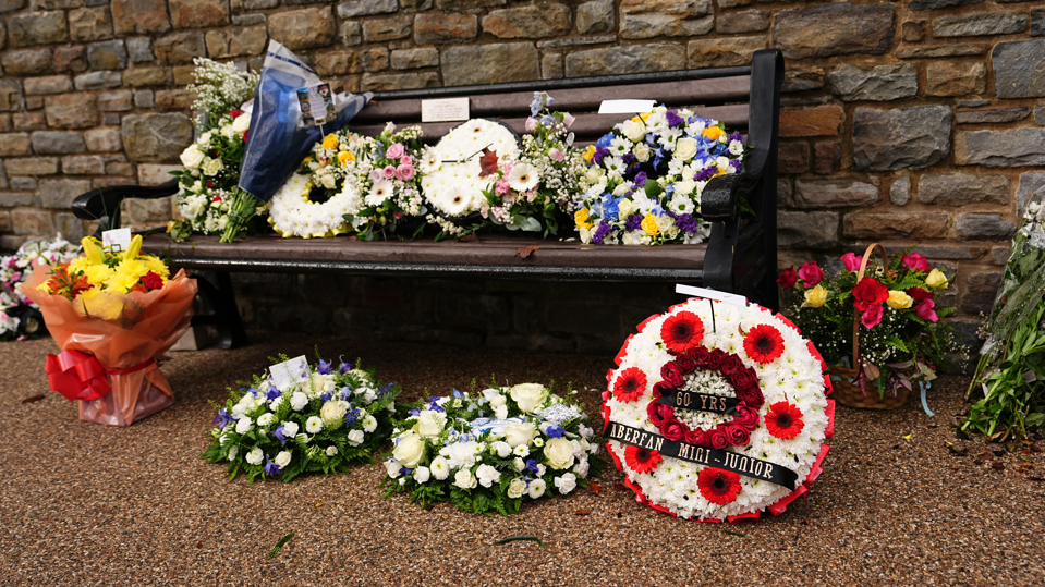 A number of flowers and wreaths have been placed beside and on a bench next to a stone wall.