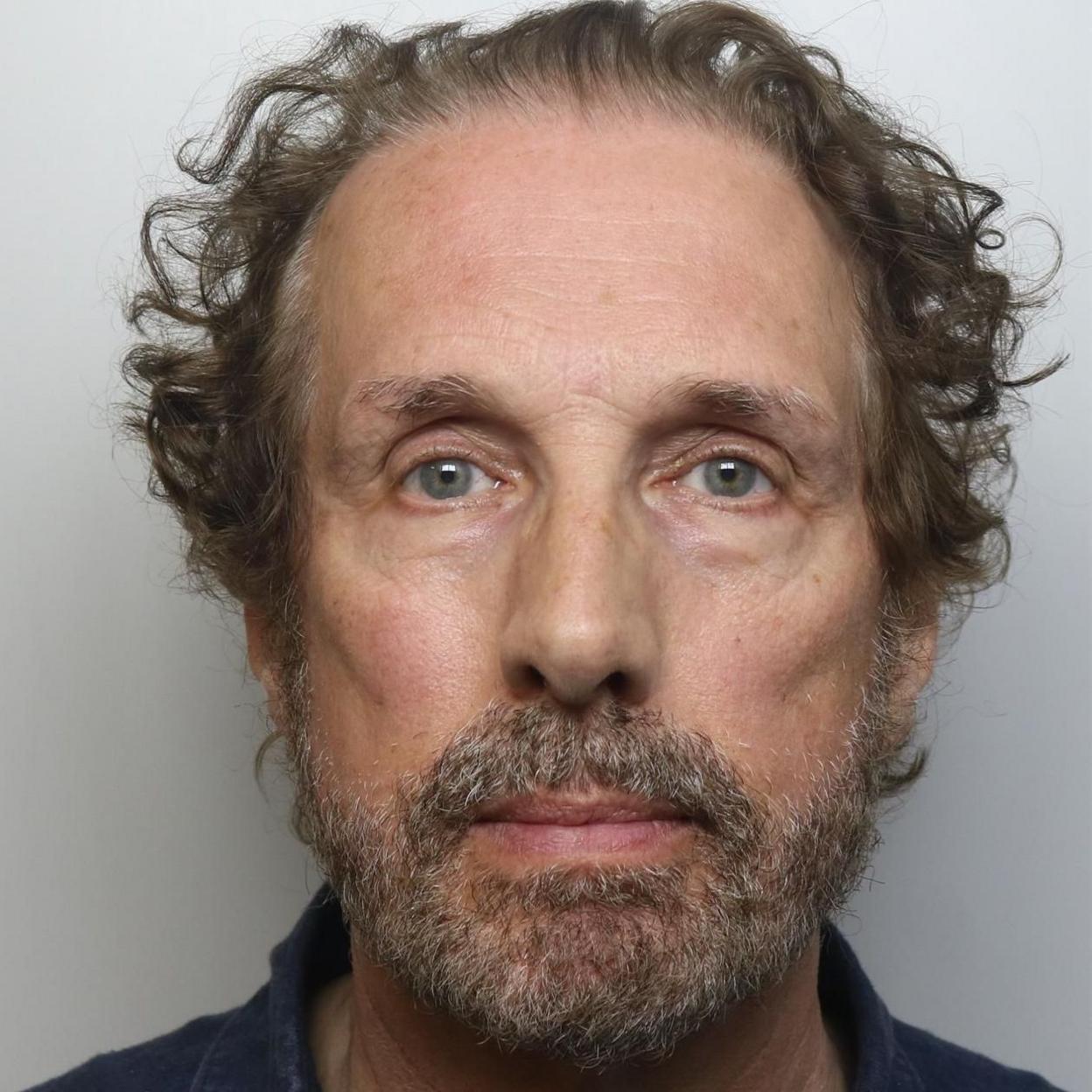 A man with blue-grey eyes, a grey beard and curly brown hair is shown in a police custody image