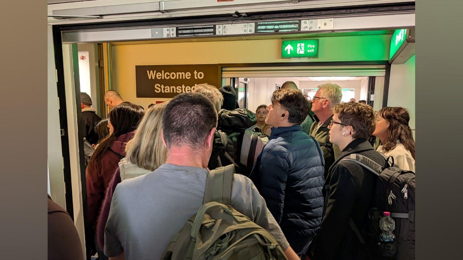 A number of people queuing in a corridor. There is a sign that reads "Welcome to Stansted" hanging on the wall.
