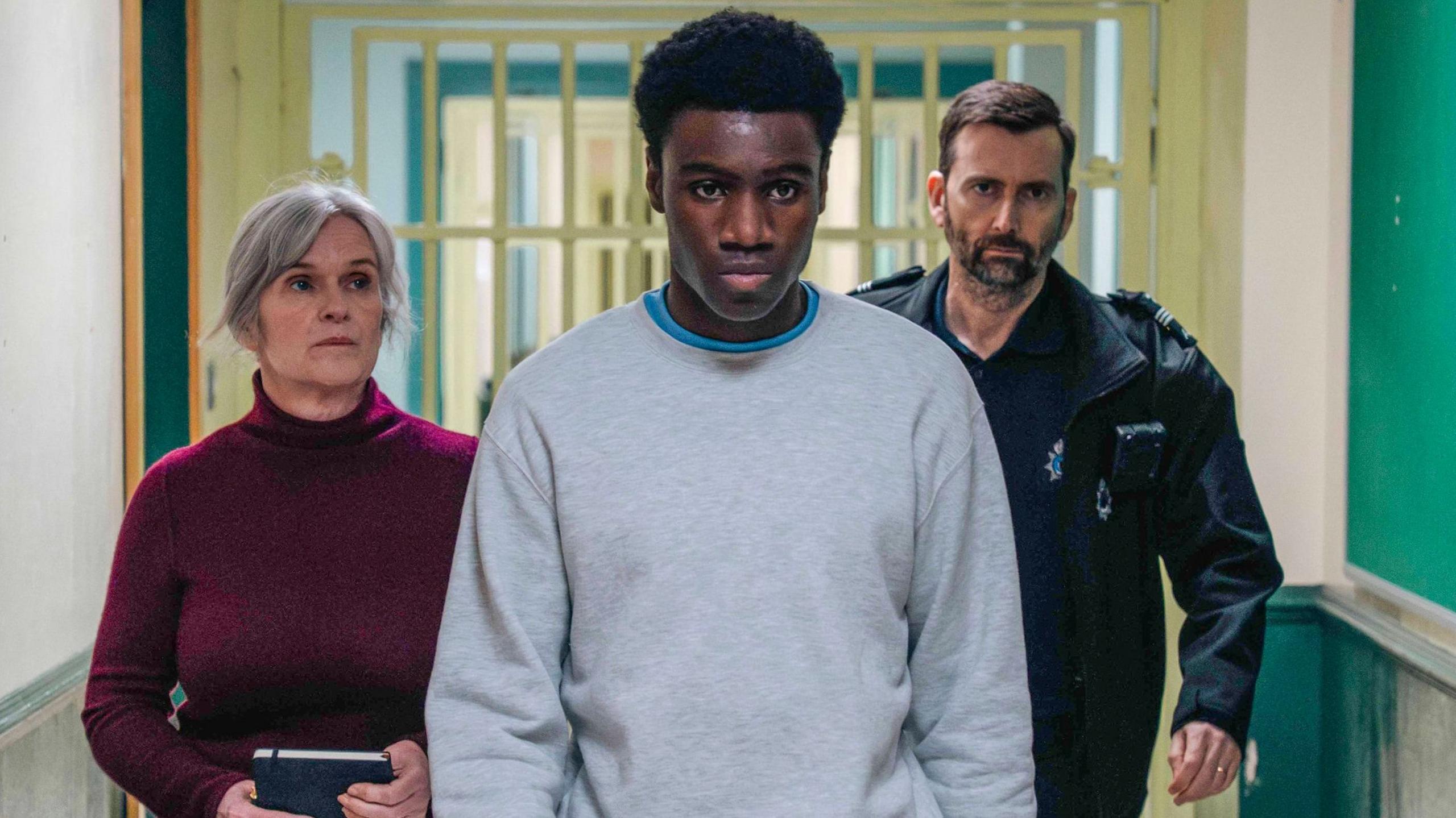 Still taken from the programme showing David Tennant and Siobhan Finneran walking along a corridor behind Chukwubuikem Molokwu who is wearing a grey tracksuit.
