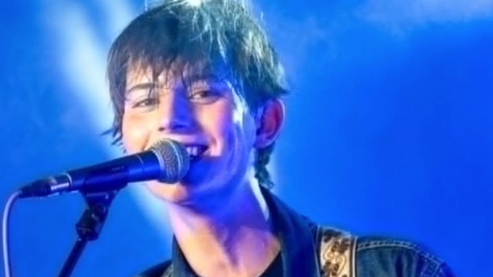 A close-up image of Kit Bottomley with short brown hair, who is smiling and singing into a microphone against a blue backdrop.