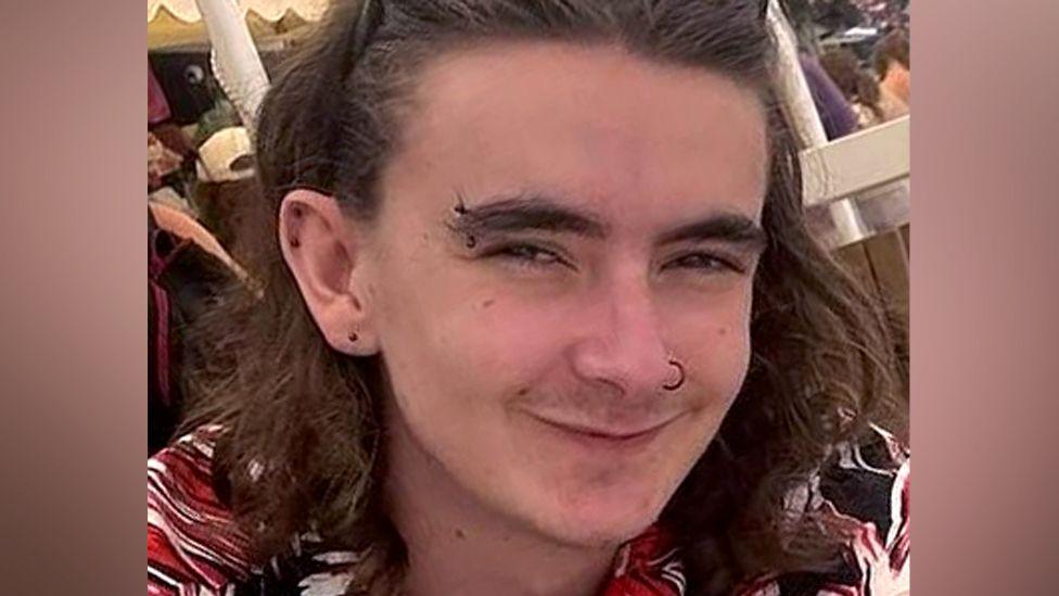 Jamie Collins wearing a black, red and white patterned shirt and sunglasses on his head. He has shoulder length brown hair and piercings in his ears, eyebrow and nose. He appears to be sitting underneath a white marquee on a sunny day.