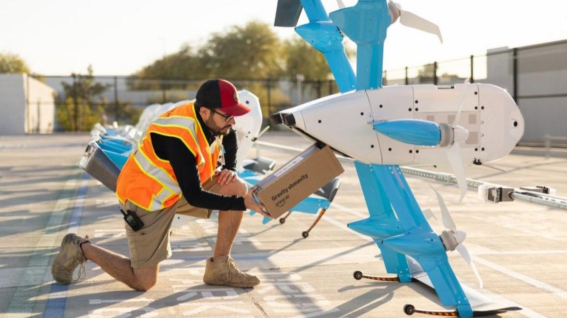 A worker kneels next to an Amazon drone and loads a package into its cargo bay. The drone is large - as big as the worker.