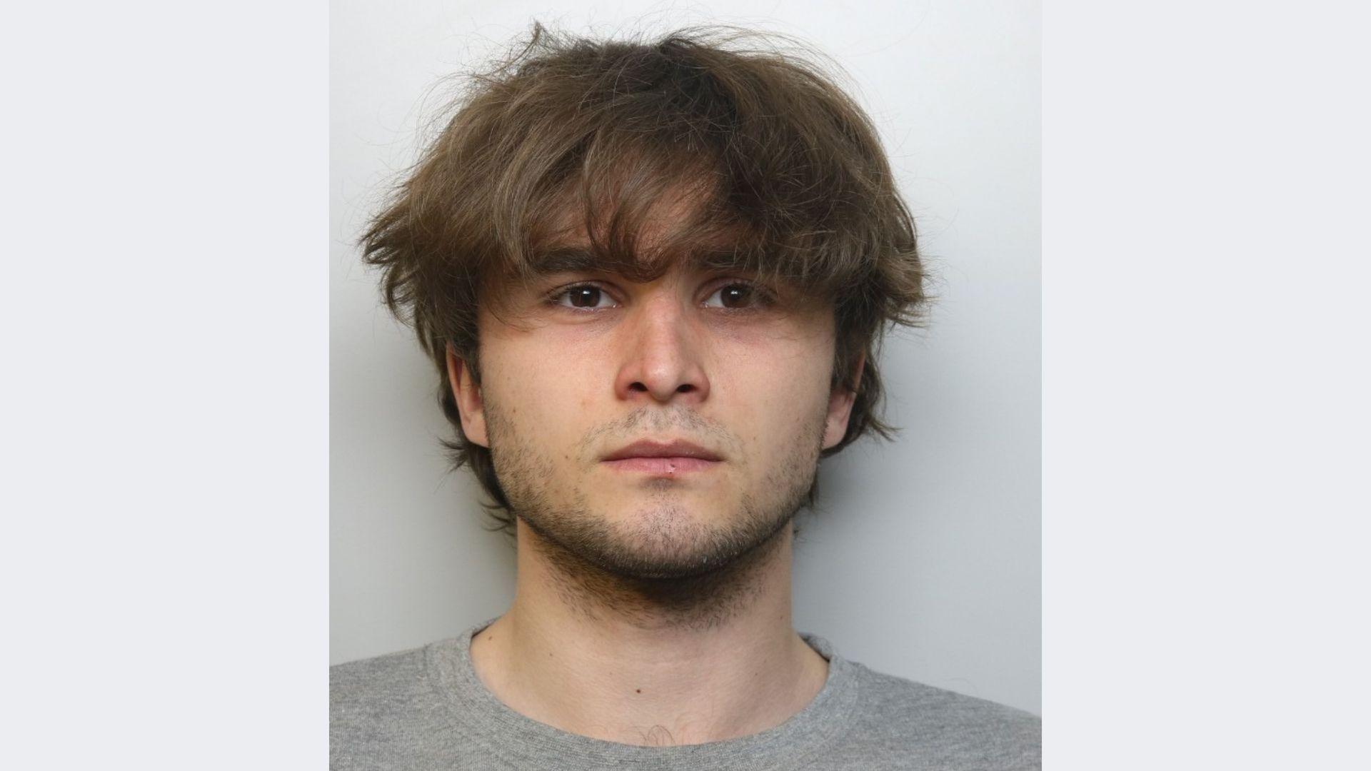 A police mugshot of Zach Coughlan wearing a grey T-shirt and standing against a grey background. He has a closely shaven beard and long messy brown hair, and is looking at the camera.
