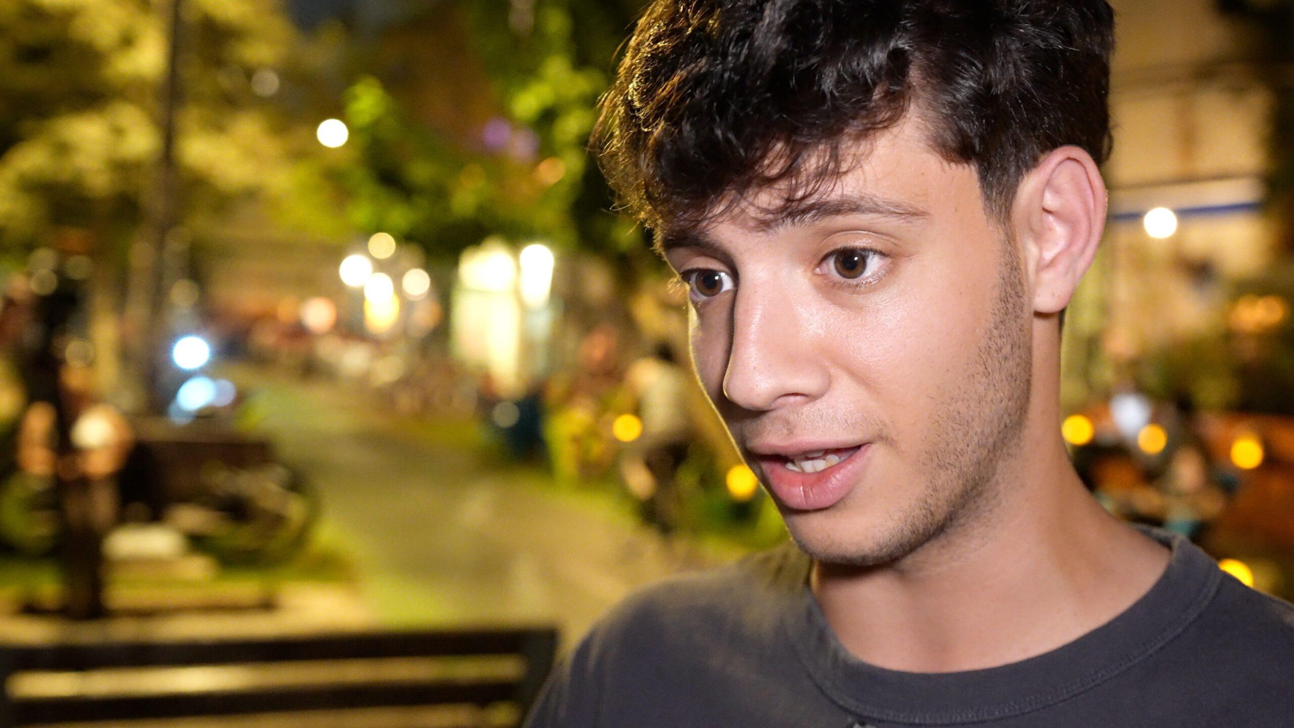 A young man with dark brown hair stands in a street at night-time, speaking to camera