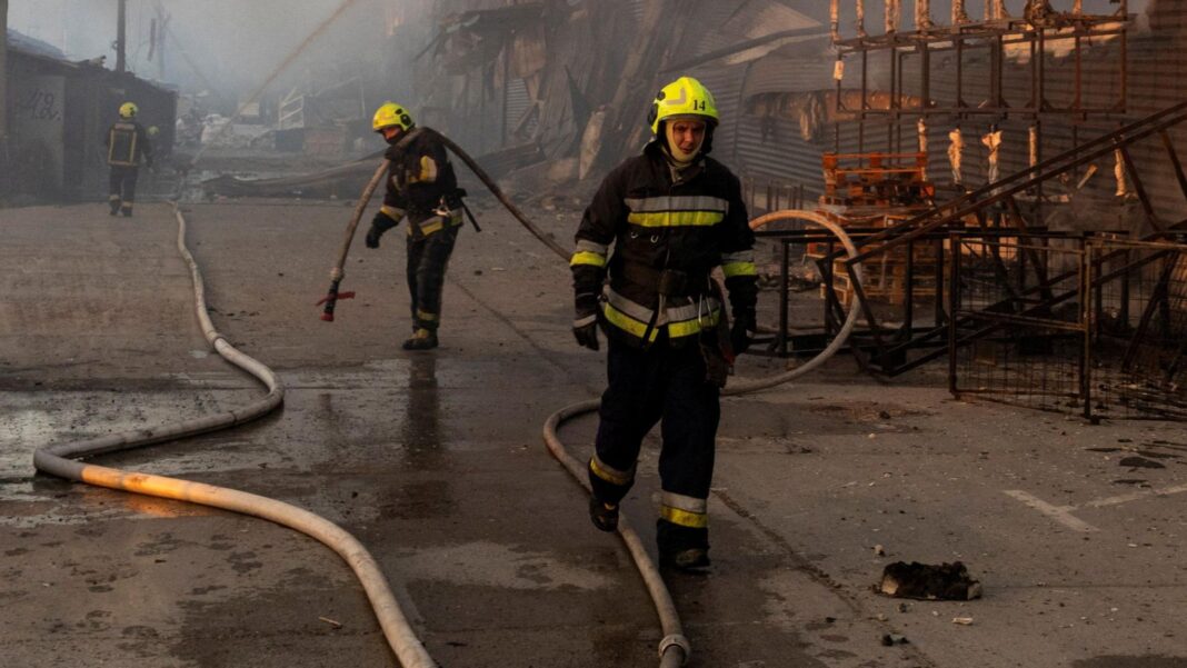 Three firefighters wearing yellow helmets work at the site of a warehouse that burned out after it was hit during a night of Russian missile attacks in Kyiv. One of the firefighters is seen holding a large hose. The destroyed remains of the warehouse can be seen in the background