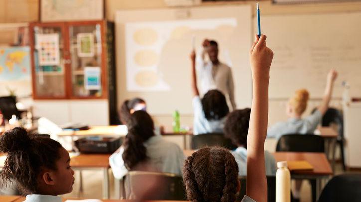A primary school classroom with pupil sitting at desks. We can see a pupil in the foreground raising her hand with a blue pencil. We can only see the backs of the pupils heads. The teacher stands at the front of the class wearing a white shirt. The photo is partially blurred
