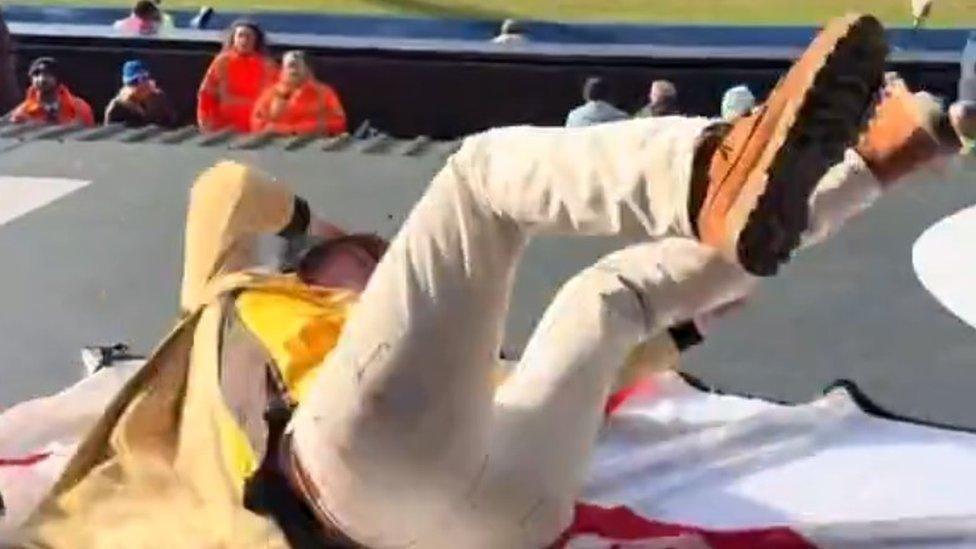 A man wearing a yellow or beige coat, and cream trousers, is on his back, appearing to fall down towards people sitting in a football stand below.
