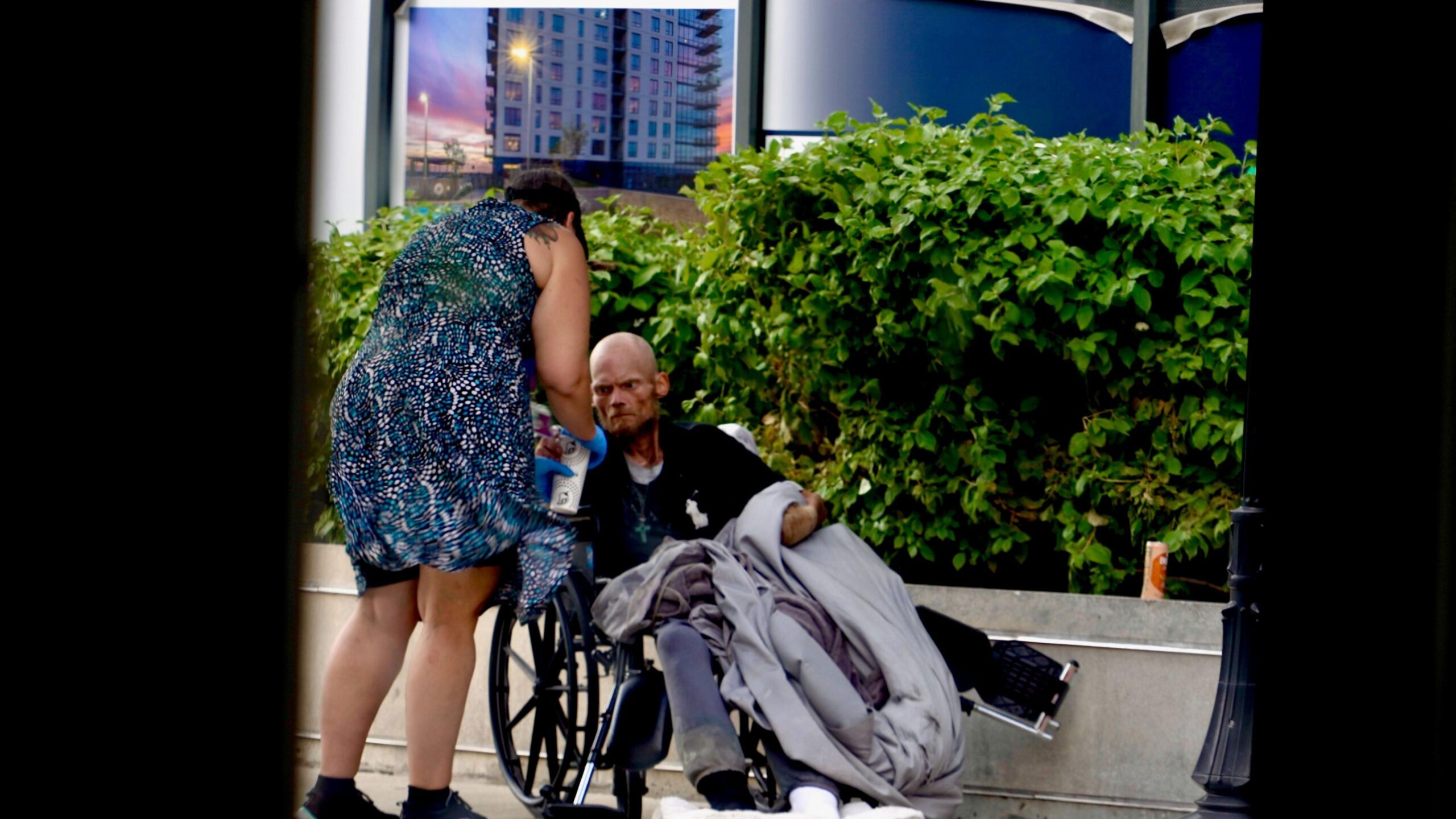 Karen, who wears a knee-length blue dress, tends to a gaunt-looking man in a wheelchair in an upscale neighbourhood