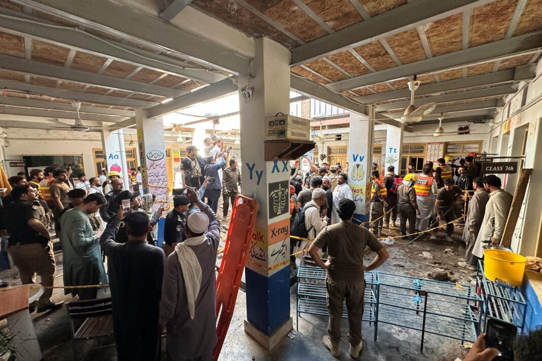 Crowds of rescue workers, police officers and local residents fill the covered courtyard of a school building as an emergency operation unfolds behind cordons. Debris cover parts of the floor, while responders in helmets and high-visibility vests work near a taped-off area. Educational murals displaying letters and vocabulary words are painted on pillars throughout the building, and several onlookers record the scene on mobile phones.