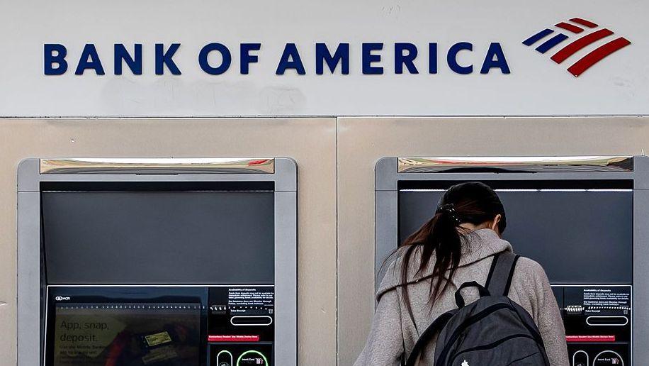 Woman stands in front of a Bank of America cash machine. She has her back to the camera and is wearing a grey coat and black rucksack