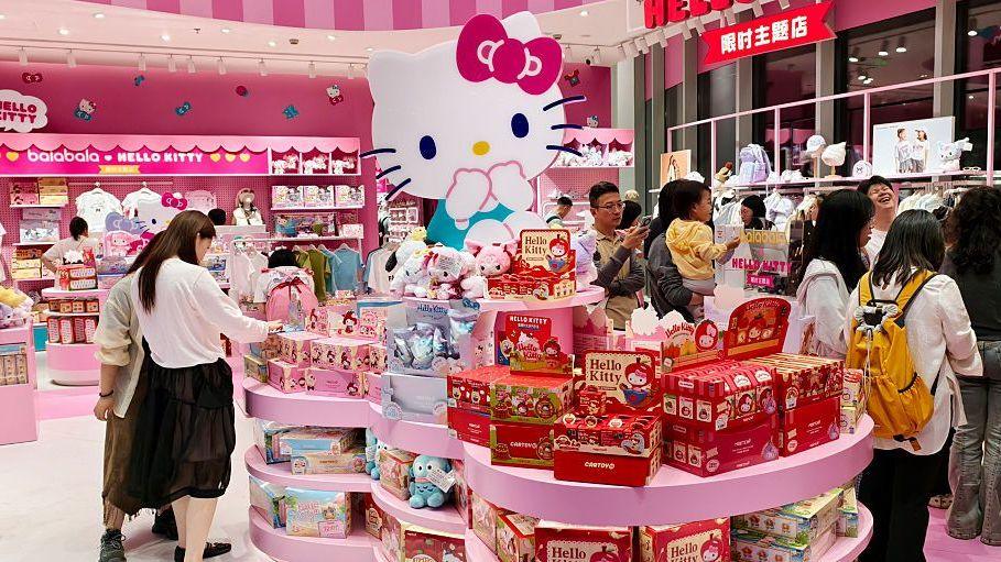 A brightly lit Hello Kitty-themed retail store filled with pink displays and merchandise. A large Hello Kitty statue stands at the centre of a circular display stacked with toys, gift sets and boxed products.