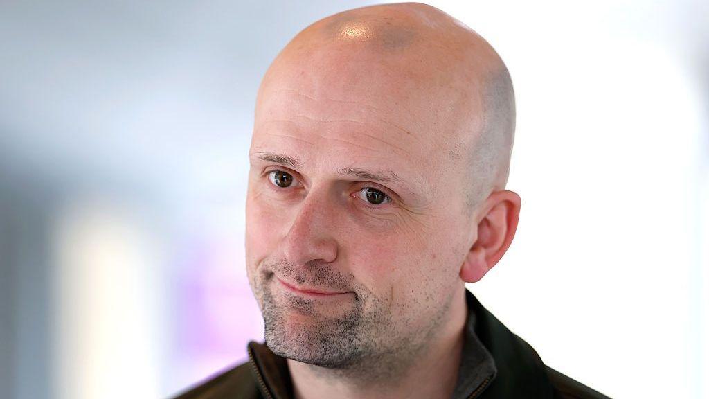 Stephen Flynn looking slightly off camera. Only his head and shoulders are visible and he appears to be wearing a dark top.