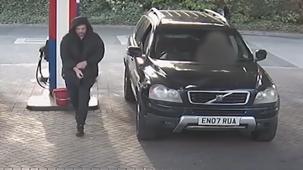 A white man wearing a black bubble coat, with the hood up, and black trousers walks across a petrol forecourt away from a black Volvo XC90 estate style car.
