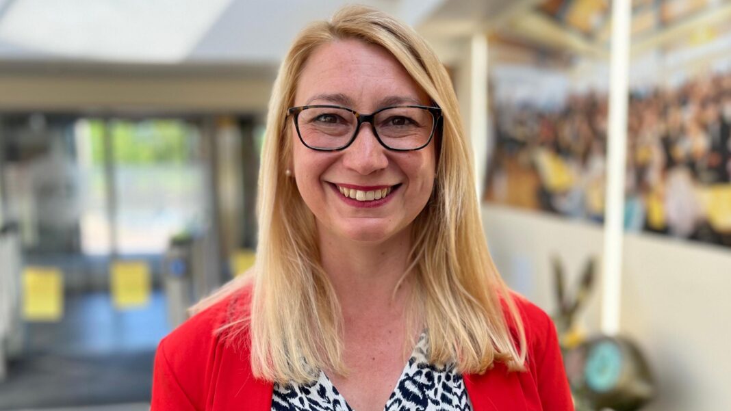 ‘Don’t axe Youth Parliament’ pleads teen member Jess Asato who is standing up while having her picture taken. She is looking directly at the camera and smiling and is wearing a white and black blouse with a red blazer over the top.