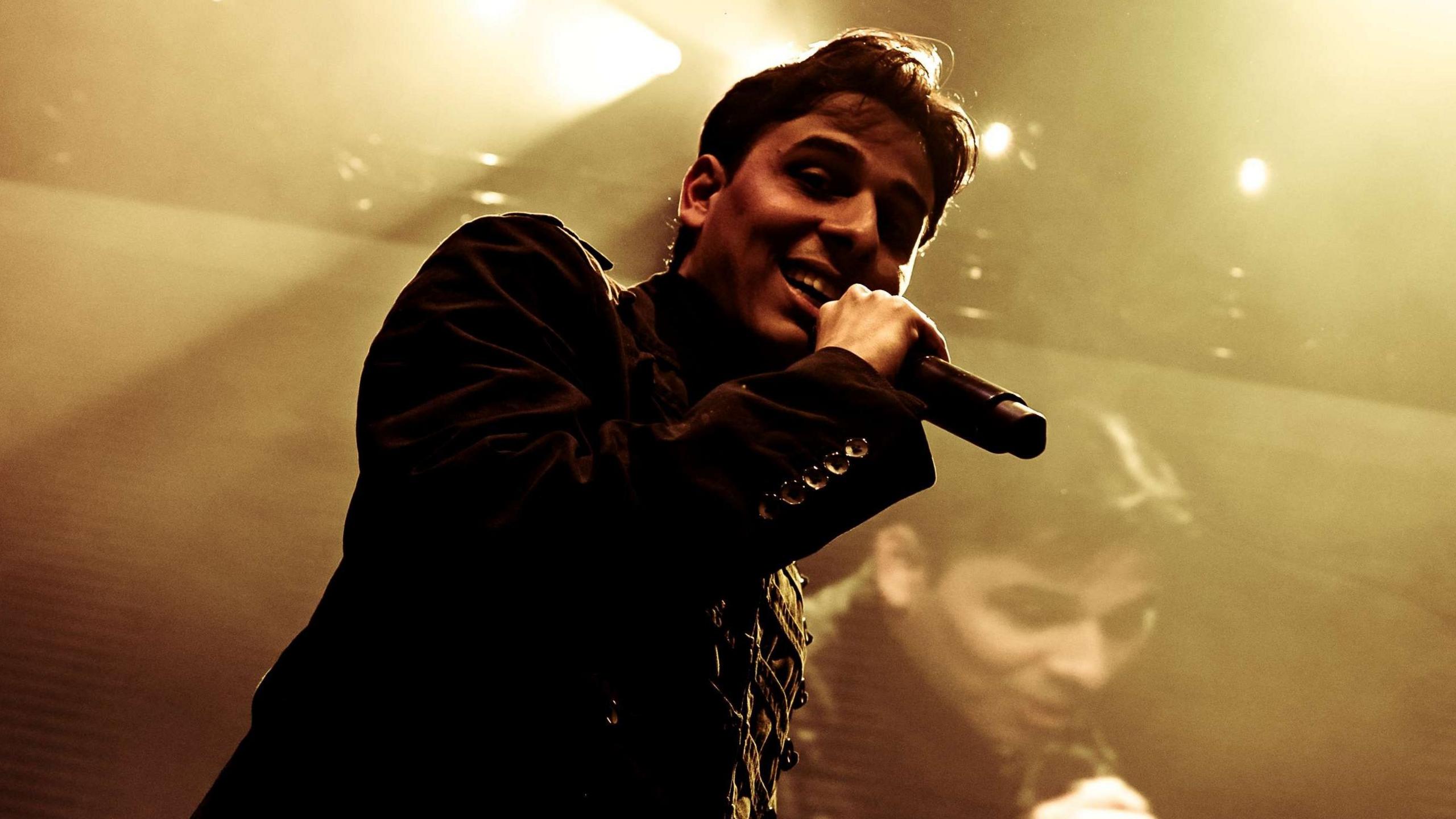 Anuv Jain performs on-stage, wearing a black buttoned up jacket and holding a microphone. He has short black hair and looks out towards the crowd, while behind him, the scene is projected on a screen