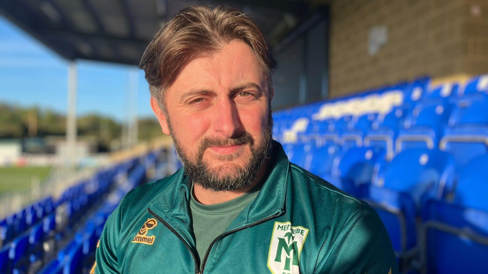 Callum Tipton sits in a football stadium stand wearing a dark green quarter-zip sports top with the “MENTRIBE FC” crest on the chest. Callum has medium length brown hair with short sides and has a short beard.