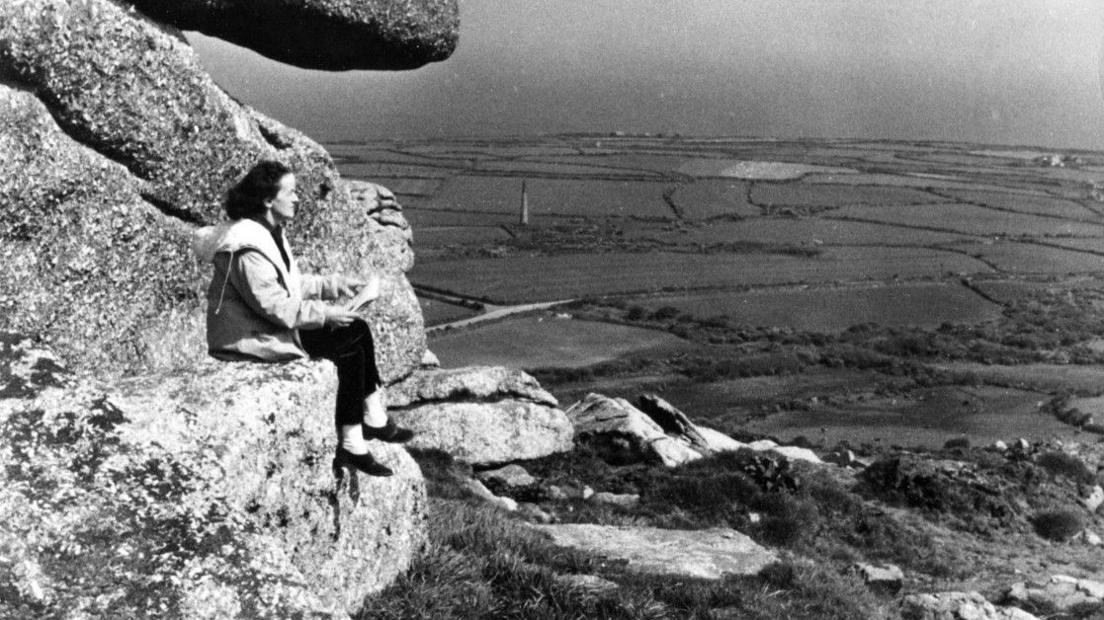 Dame Barbara Hepworth is sitting on a huge piece of stone. She has a sketchbook in her hands and is looking across fields. She is wearing dark trousers and a pale jacket in this black and white still from a BBC film.
