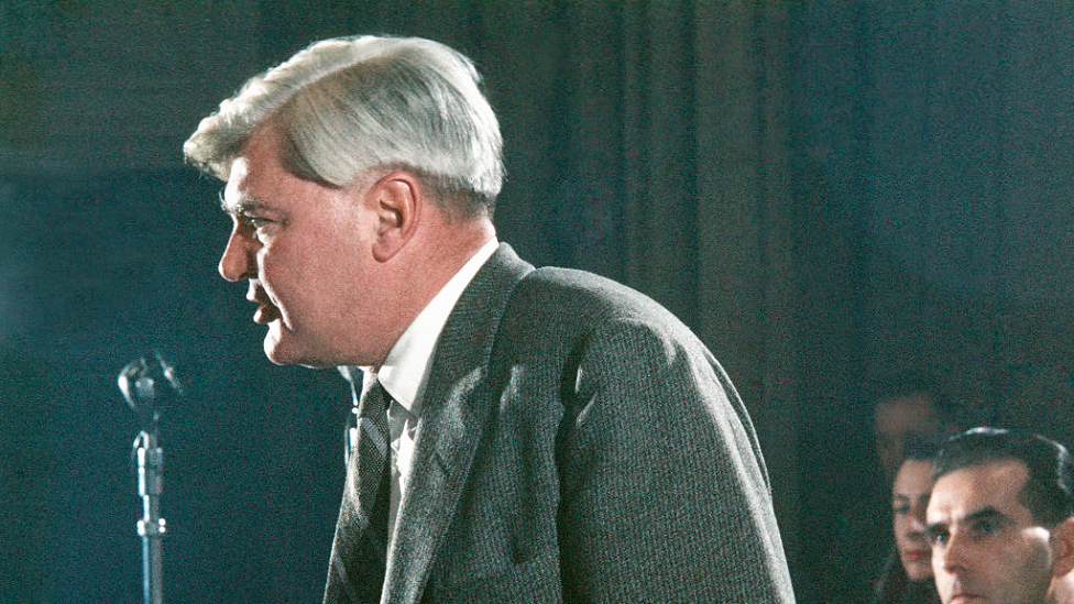 A colour photo of Aneurin Bevan in 1958 taken from the side as he is speaking in front of a microphone. He is facing towards the left of the picture. He wears a dark grey pinstripe suit, white shirt and grey striped tie. His white hair is parted on the left side and is thick. Behind him three people can be seen sitting in a row, looking at him speaking.