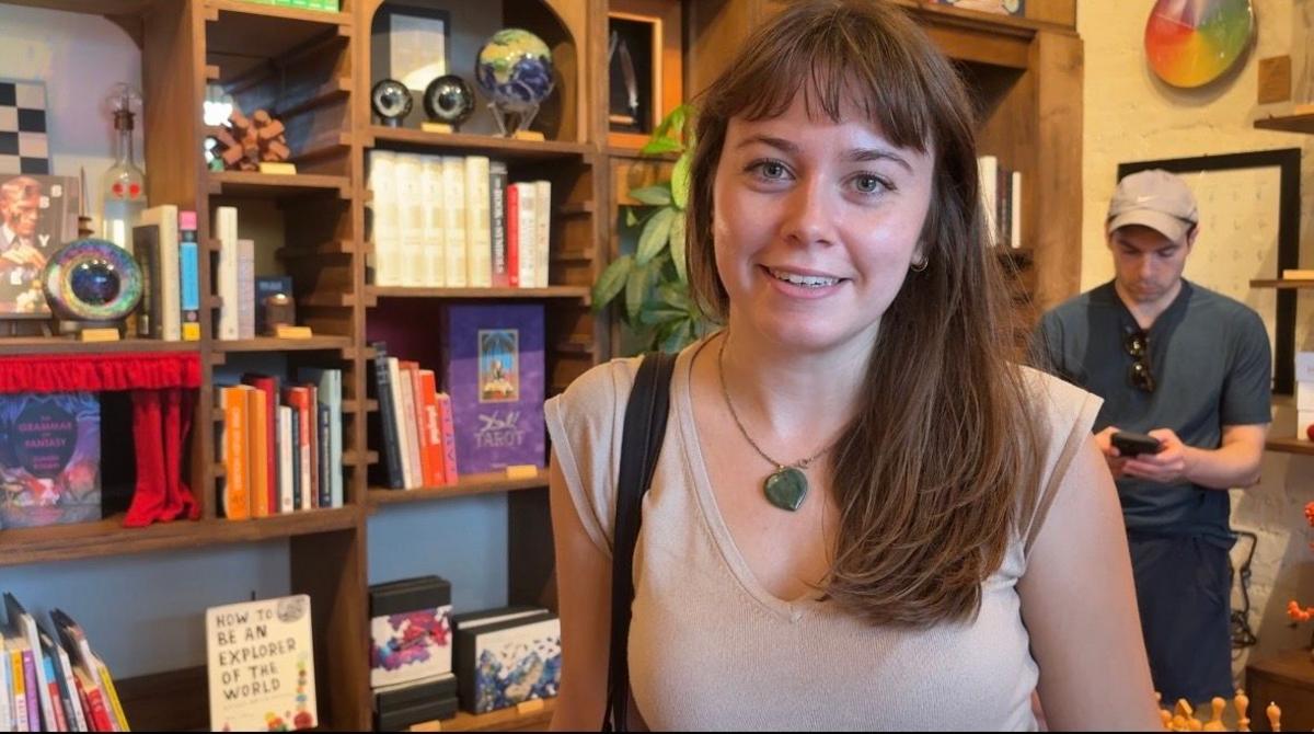 A woman with brown hair and wearing a light coloured top and necklace with a green pendant smiles at the camera. Behind her are bookshelves filled with books and other items and a man in a cap looks at his phone.