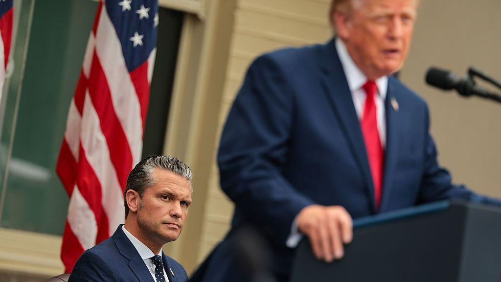 Pete Hegseth seated while President Trump stands at the lectern in a blue suit and red tie
